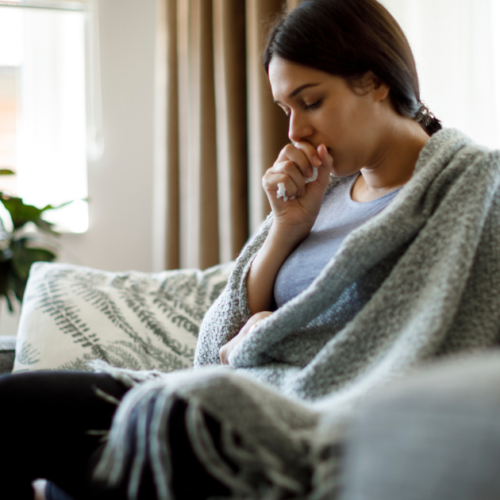 A young woman sits on a sofa wrapped In a blanket, holding a tissue in their hand